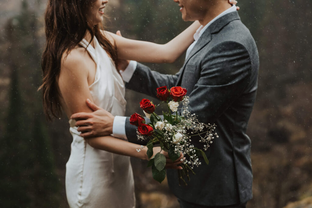 Elopement couple smiling in the rain and the bride is holding red flowers in her hand.